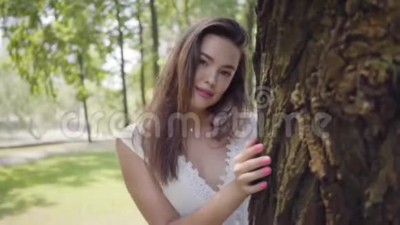 一个可爱的小女孩长着一头深色长发穿着一件白色的夏装站在一棵树旁边视频的预览图