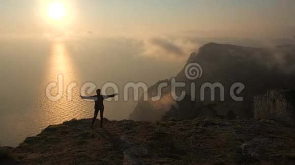 一位年轻女子伸出双臂从一座高山望着海面上美丽的日落视频的预览图