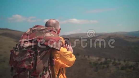 一个穿着黄色夹克和眼镜的人站在山上欣赏风景在电话里拍照视频的预览图