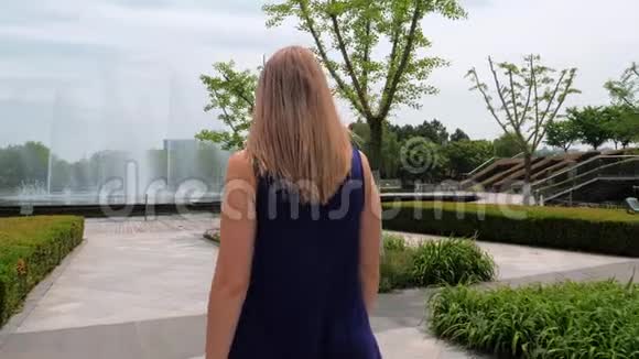 斯塔迪加姆的后景拍摄了一个穿着蓝色裙子的漂亮女孩在公园里走到喷泉前视频的预览图