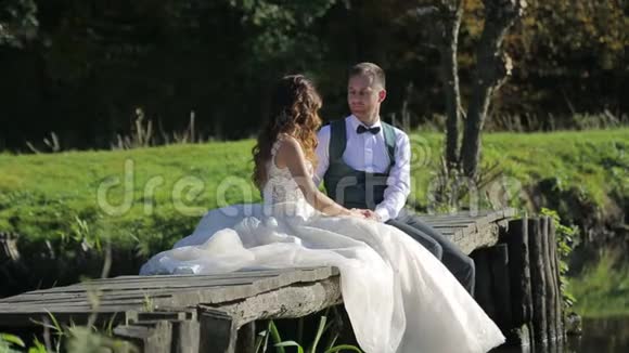 新娘坐在河边的一座木桥上一对幸福的家庭夫妇花时间在公园里视频的预览图