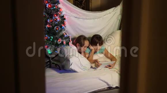在装饰着五颜六色圣诞树的房间里玩平板电脑的姐妹们家庭圣诞节的概念儿童视频的预览图