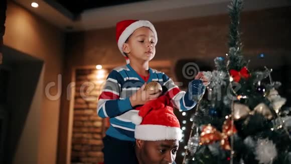 一个孩子正在用闪闪发光的球装饰一棵圣诞树坐在他父亲肩膀上视频的预览图