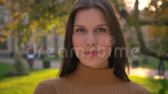 在绿色公园的背景下年轻的黑发女孩观看镜头调情和微笑的特写肖像视频的预览图