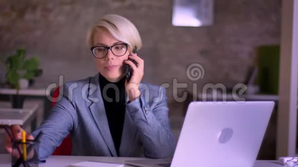 一幅中年金发戴眼镜的短发女商人在办公室里高兴地用手机说话的肖像视频的预览图