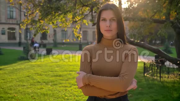 年轻的黑发女孩在绿色公园的背景下平静地看着镜头手臂交叉视频的预览图