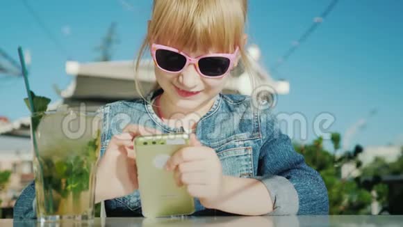 戴着粉红色眼镜的有趣女孩在智能手机上玩在咖啡馆的夏日公寓里视频的预览图