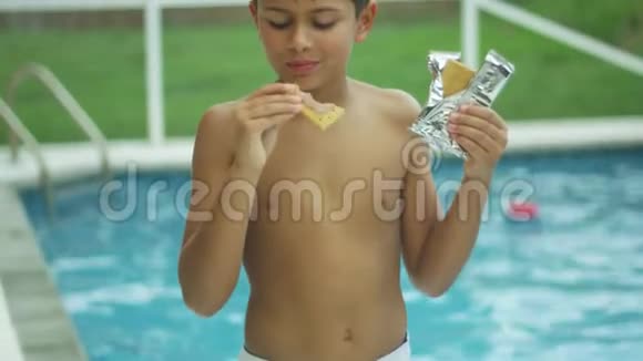 一个小男孩在游泳池前微笑着吃东西的慢动作视频的预览图