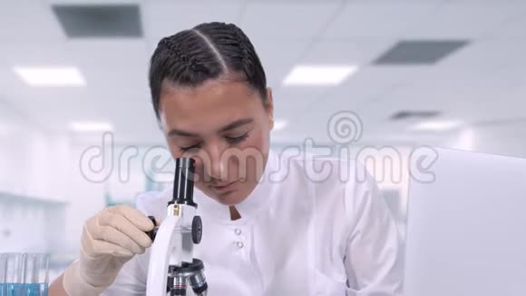 一位女性医学专业人员在显微镜下观察生物样本并在坐着时将数据写入笔记本电脑视频的预览图