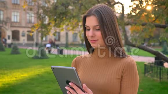 年轻白种人女孩使用平板电脑的肖像并在绿色公园的背景上显示其显示到相机上视频的预览图