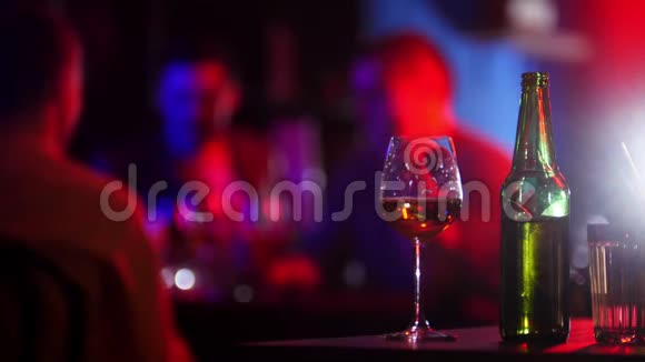 带霓虹灯的酒吧一个年轻人拿着绿色的啤酒瓶坐在他的朋友旁边干杯视频的预览图
