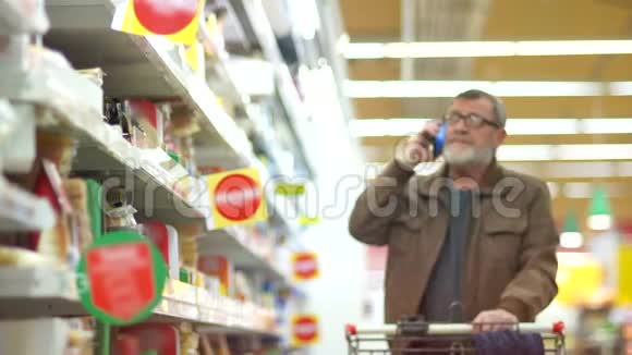 在乳品部超市的成熟男人养老金领取者在电话中交谈选择奶酪放在货架上视频的预览图