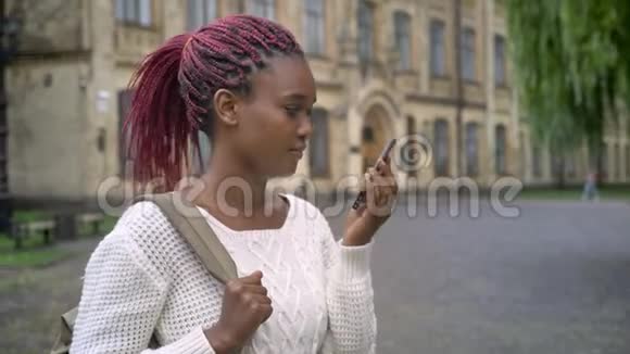 年轻迷人的非洲女学生打电话拿着背包站在大学附近的公园里视频的预览图