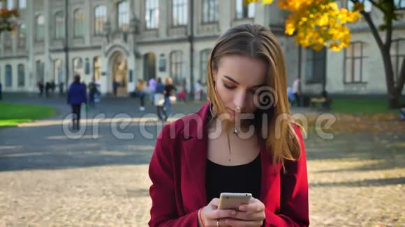 年轻的女学生站在电话里发短信生气然后微笑在一个阳光明媚的日子里走出大学视频的预览图
