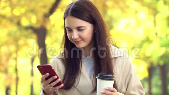 在公园里穿外套或壕沟的女人带着咖啡和手机视频的预览图