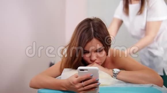 一位年轻女子接受放松的背部按摩坐在手机里看着屏幕视频的预览图