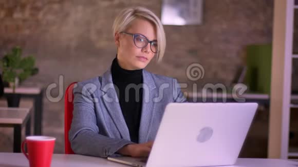 戴着眼镜的金发短发女商人的肖像专心地看着笔记本电脑喝着咖啡视频的预览图