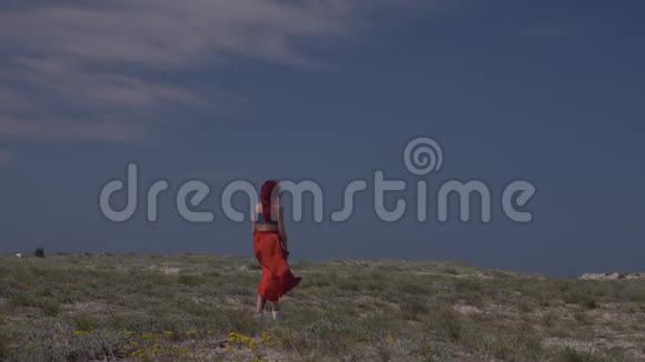 一位身穿红色裙子的红发年轻女子在蓝天上行走的后景视频的预览图