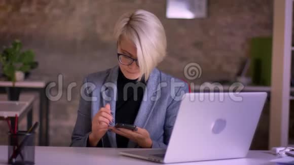 中年短发女商人戴着眼镜在办公室的笔记本电脑前用智能手机工作的肖像视频的预览图