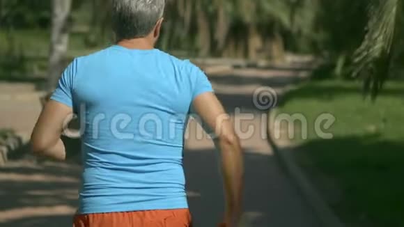 穿着蓝色t恤的年轻灰发男子沿着热带公园路奔跑特写慢镜头视频的预览图