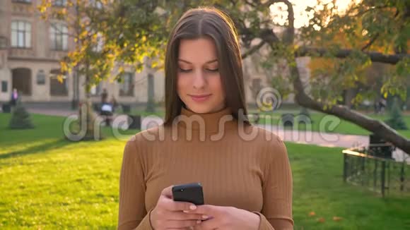可爱的高加索女孩冲浪和聊天使用智能手机并改变情绪在绿色公园的背景视频的预览图