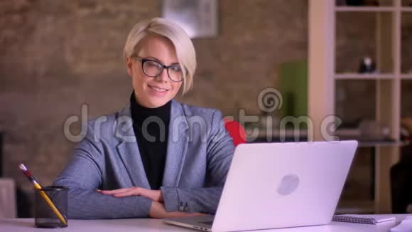 戴着眼镜和双手的金发短发女商人的肖像交叉在桌子上微笑着进入办公室的镜头视频的预览图