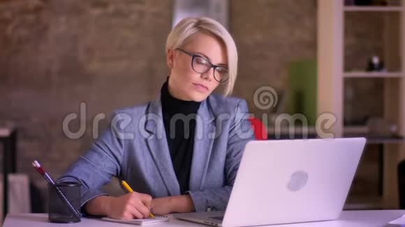 一位戴眼镜的金发短发女商人的肖像在办公室的笔记本上认真地做笔记视频的预览图