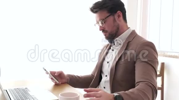 英俊的年轻商人在办公室的笔记本电脑前喝咖啡时使用他的智能手机视频的预览图