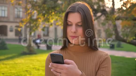 美丽的黑发女孩的肖像滚动饲料使用智能手机与各种情绪在绿色公园的背景视频的预览图