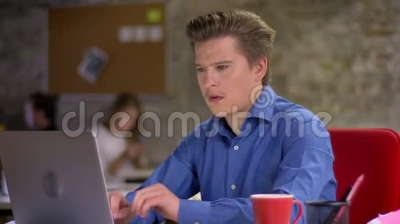 中年金发白种人商人在办公室里用笔记本电脑打字喝咖啡对着镜头微笑的肖像视频的预览图