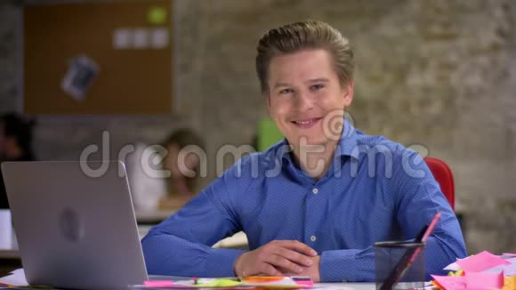 中年金发商人在笔记本电脑前的肖像在办公室里对着镜头微笑和眨眼视频的预览图