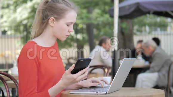 坐在咖啡厅露台上使用智能手机和笔记本电脑的年轻女子视频的预览图