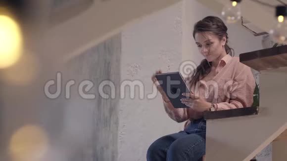穿着粉红色衬衫和牛仔裤的美丽微笑的女人坐在家里的木楼梯上打字视频的预览图