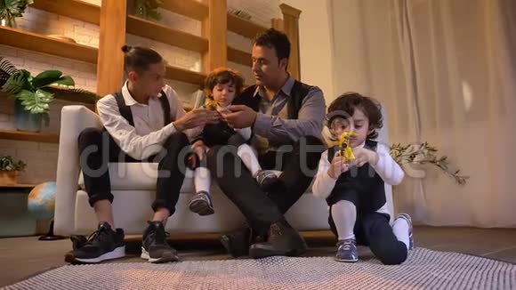 阿拉伯父亲和他的双胞胎儿子和十几岁的孩子坐在沙发上在客厅里玩玩具的肖像视频的预览图