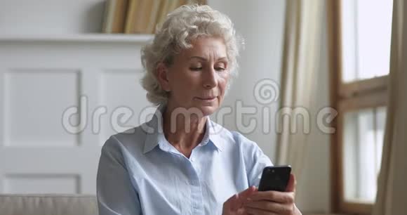 微笑的老年妇女使用智能手机坐在家里的沙发上视频的预览图