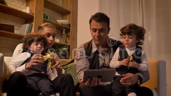 阿拉伯语的父亲和他十几岁的儿子坐在沙发上两个卷发男孩在客厅里看着平板电脑视频的预览图