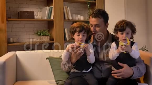 阿拉伯中年父亲坐在沙发上的肖像他的双胞胎儿子在客厅里玩玩具机器人视频的预览图