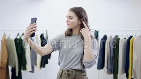年轻漂亮的女人在智能手机上自拍在服装室里玩头发视频的预览图
