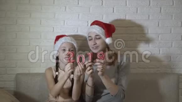 两个戴着圣诞帽子的小女孩拿着2019年的红色数字坐在沙发上跳舞微笑着视频的预览图