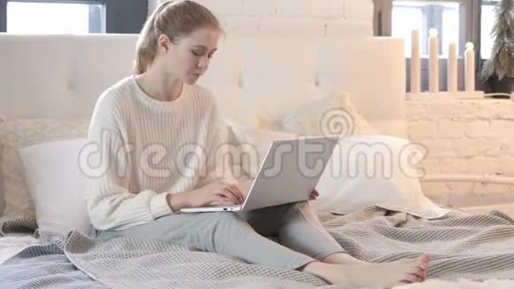 颈部疼痛的疲惫年轻女子在床上做手提电脑视频的预览图
