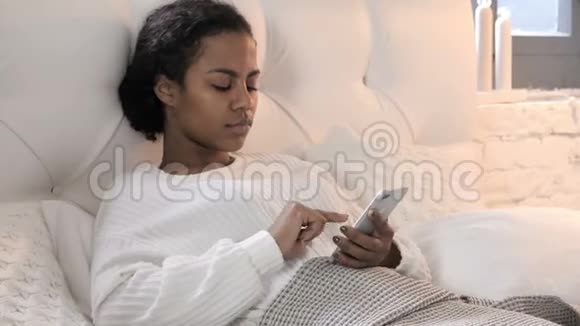 年轻的非洲女性在床上放松时使用智能手机视频的预览图