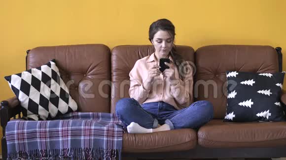 穿着粉红色衬衫和牛仔裤的年轻漂亮女人坐在棕色沙发上抱着枕头在智能手机上打字视频的预览图