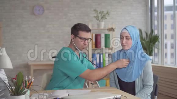 年轻的男医生用听诊器听一个戴着头巾的年轻穆斯林妇女的心跳视频的预览图