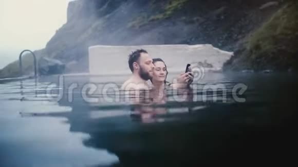 年轻漂亮的夫妇在冰岛的温泉里游泳旅行男女在智能手机上自拍照片视频的预览图