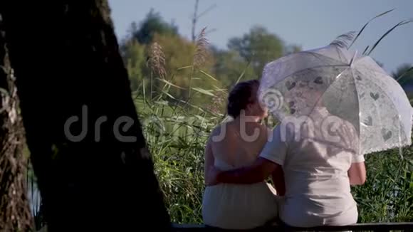 女人和她的爱人拿着白伞坐在河边的长凳上视频的预览图