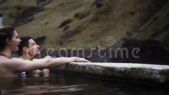 年轻的幸福夫妇在冰岛的温泉里游泳旅行的男人和女人在山上欣赏风景视频的预览图