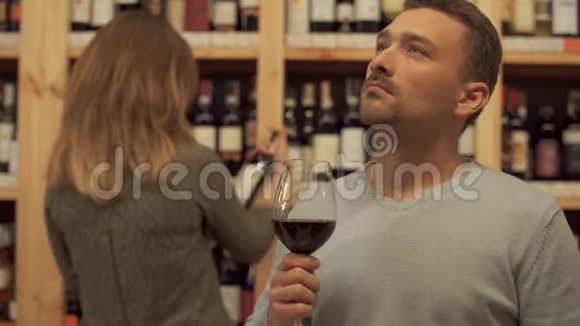 在一家酒类商店里拿着酒杯的帅哥背景中的女人正在选择一个酒瓶视频的预览图