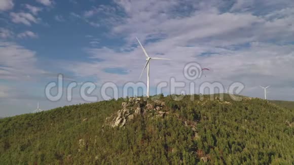 在一座长满绿树的山顶上跳伞者在风力发电机附近飞行视频的预览图