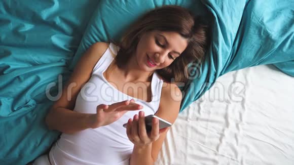 早上躺在床上用智能手机应用程序晒黑的女人的俯视图视频的预览图
