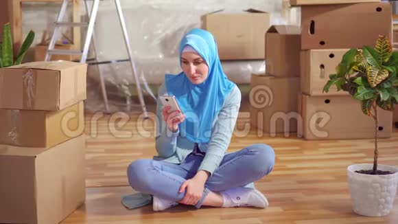 戴着头巾的年轻穆斯林妇女使用智能手机坐在一间现代化公寓的盒子旁边的地板上视频的预览图